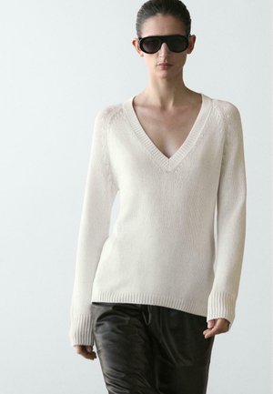 Kvinde iført oversized sorte solbriller, en cremefarvet strikket trøje med V-hals og sorte læderbukser foran en ensartet lys baggrund.