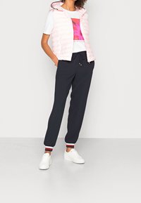 Chaleco acolchado rosa claro, camiseta gráfica blanca y pantalones de chándal azul marino con puños a rayas. Combinado con zapatillas blancas, con parte inferior elástica.