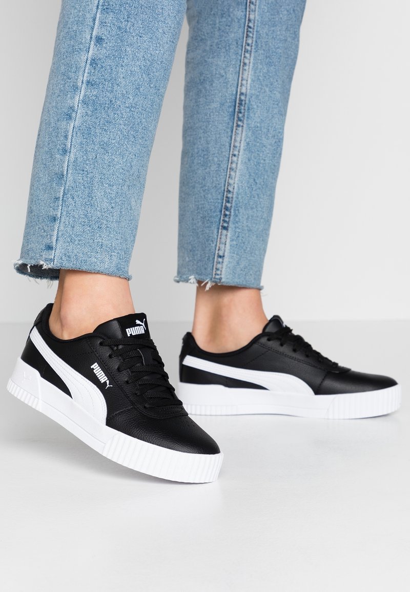 Svarta lädersneakers har en vit rand och en texturerad sula. De matchas med ljusblå fransade jeans, vilket betonar moderna designelement.
