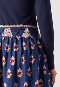 Robe navy avec une taille smockée et des motifs floraux contrastants en rouge et blanc. Manches longues et tissu doux et fluide.