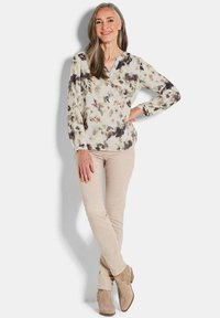 Blumige Bluse in Creme mit dunklen Akzenten, geknöpftem Ausschnitt und langen Ärmeln; kombiniert mit beigefarbenen, figurbetonten Hosen und taupefarbene Ankle Boots.