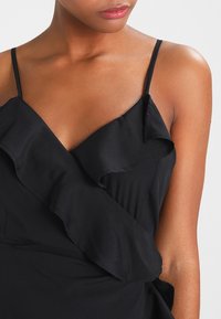 Svart ärmlös klänning med tunna band, med en volanginslaget design och mjuk textur. Närbild av övre delen som lyfter fram detaljer.