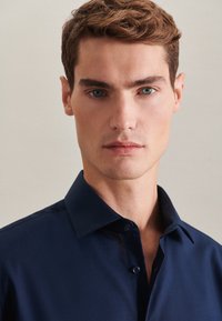 Camicia blu navy con colletto classico e texture liscia. Presenta un design aderente e una leggera lucentezza, con bottoni davanti.