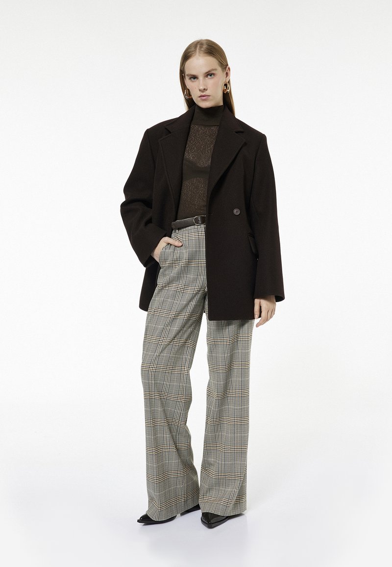 Brązowa, oversize'owa marynarka, dopasowana, prześwitująca bluzka, szerokie spodnie w kratę w beżowo-czarnym wzorze, noszone z czarnymi, szpiczastymi szpilkami.