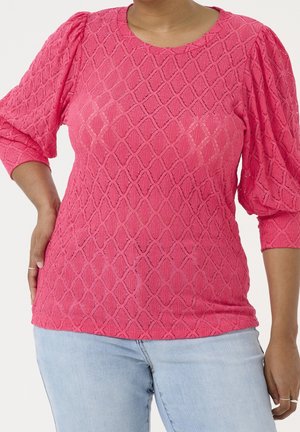 Frau trägt eine leuchtend pinke, strukturierte Bluse mit Diamantmuster und Puffärmeln, kombiniert mit hellblauen Jeans, steht mit einer Hand in der Hüfte.