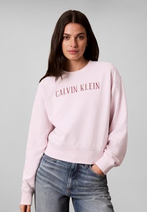 Kobieta z długimi ciemnymi włosami, ubrana w jasnoróżową bluzę Calvin Klein i niebieskie dżinsy, stojąca z jedną ręką w kieszeni na szarym tle.