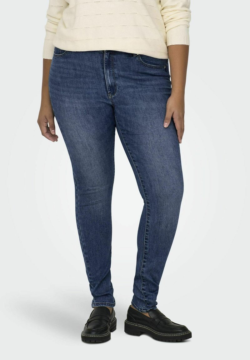 Dark blue skinny jeans made of denim, featuring a classic five-pocket design and slight fading. Worn with a cream sweater and black loafers.