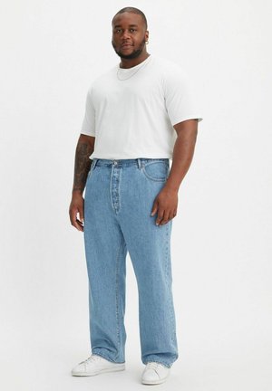 Witte korte mouwen t-shirt, lichtblauwe rechtlopende jeans en witte sneakers. Persoon staat zelfverzekerd met de handen in de zakken.