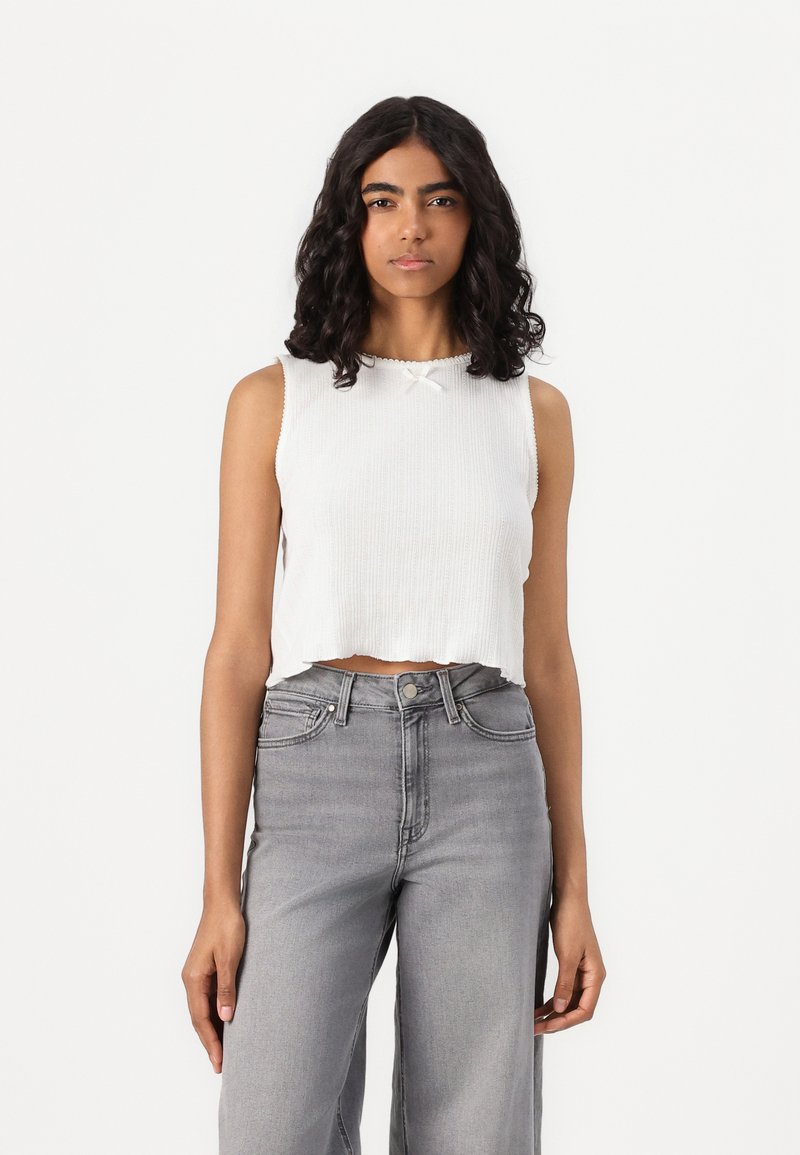 Top bianco a maglia tagliato, con una texture a coste, dotato di scollatura tonda e dettaglio a fiocco, abbinato a jeans grigi a gamba larga e a vita alta.