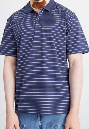 Mørkeblå polo-shirt med hvide vandrette striber, korte ærmer og krave. Har en enkelt knaplukning ved kraven.