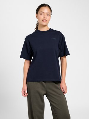 Ung kvinde med mørkt hår i hestehale iført en navyblå kortærmet t-shirt og olivengrønne bukser, stående foran en ensfarvet baggrund.