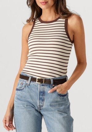 Vrouw draagt een mouwloos beige en bruin gestreept topje, ingestopt in een hoog getailleerde blauwe spijkerbroek met een donkerbruine riem, één hand in de zak.