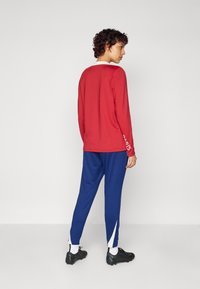 Chemise rouge à manches longues avec un col blanc et des inscriptions sur la manche, associée à un pantalon bleu avec des accents blancs sur les côtés et des chaussures à crampons noires.