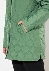 Pletená zelená bunda se strukturovaným designem, má přední zip a boční kapsy; látka vypadá hladce s jemným leskem.