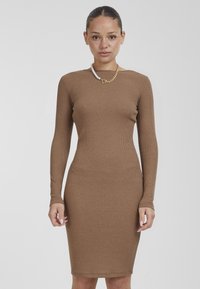 Abito lungo a maniche lunghe color marrone con silhouette aderente, orlo fino al ginocchio e scollatura rotonda. Gli accessori notevoli includono una collana di perle e una catena d'oro.