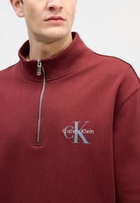 Μπορντό φούτερ με φερμουάρ και ψηλό γιακά, με ασημένιο φερμουάρ και λογότυπο "Calvin Klein" σε ανοιχτό μπλε, φτιαγμένο από μαλακό ύφασμα.