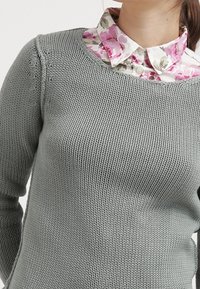 Grå stickad tröja med rund halsringning, med en blommig skjorta under i rosa, vitt och grönt mönster.