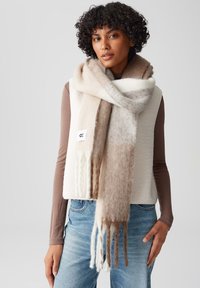Sciarpa di lana morbida a righe crema e marroni con estremità frangiate, indossata sopra un gilet beige e abbinata a jeans azzurri chiari.