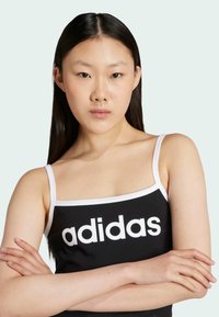 Černý top bez rukávů s bílými akcenty, na kterém je výrazné bílé logo "adidas". Hladká látka a tenké ramínka, modelka s překříženýma rukama.