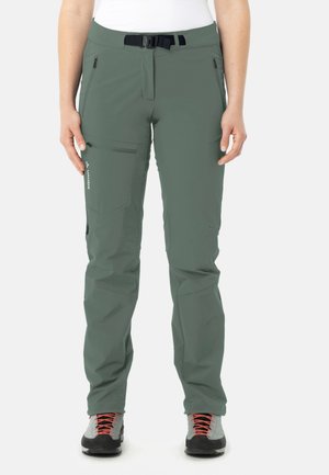 Person trägt grüne Outdoorhose mit Reißverschlusstaschen und schwarzem Gürtel, kombiniert mit grauen Wanderschuhen mit orangen Schnürsenkeln.