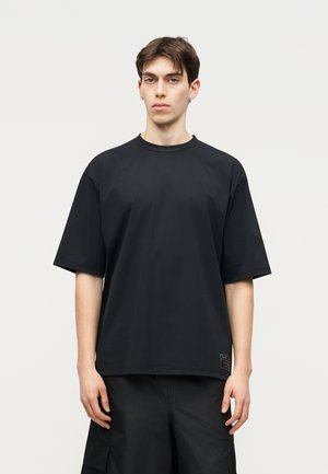 Jaunas vyras su trumpais rudais plaukais, dėvintis per didelį juodą marškinėlį ir juodus kargo šortus, stovintis priešais paprastą šviesią sieną.