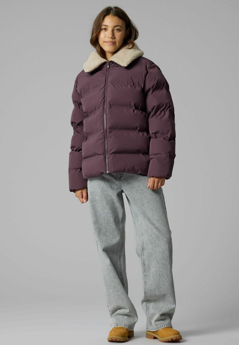 Jeune femme debout portant une doudoune violette avec un col en polaire crème, un jean bleu clair et des bottes beige sur un fond gris.
