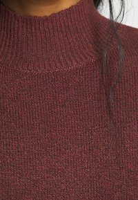 Gros plan d'une personne portant un pull à col roulé côtelé couleur bordeaux avec une mèche lâche de cheveux foncés sur le côté droit.