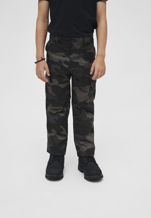 Barn iført sorte og grå camouflage cargobukser med sorte støvler og en sort kortærmet skjorte, stående mod en ensfarvet baggrund.