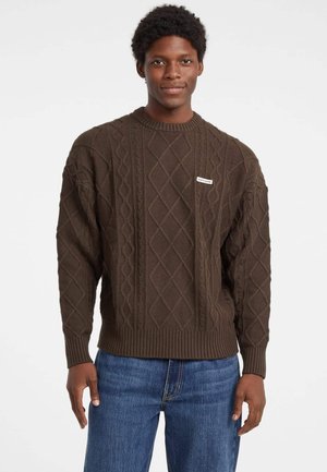 Maglione marrone a treccia con collo e polsini a costine. Presenta una piccola etichetta sul davanti. Abbinato a jeans in denim blu.