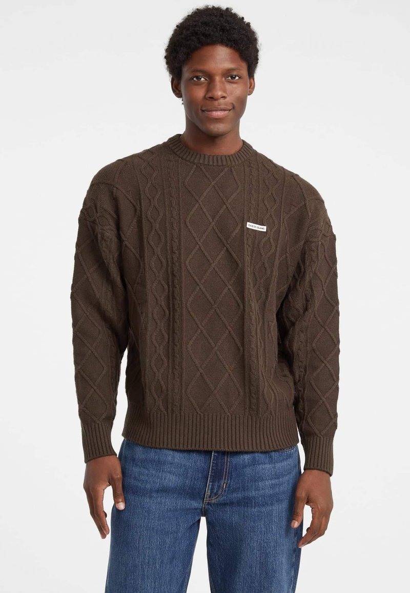Brauner Kabelstrickpullover mit geripptem Ausschnitt und Bündchen. Enthält ein kleines Etikett auf der Vorderseite. Kombiniert mit blauen Jeans.