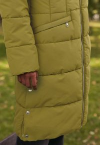 Maslinasto zeleni puffer kaput s horizontalnim šavovima, zatvaračem sprijeda, bočnim džepovima i dugmadi na rubu. Mekana, qulted tekstura.