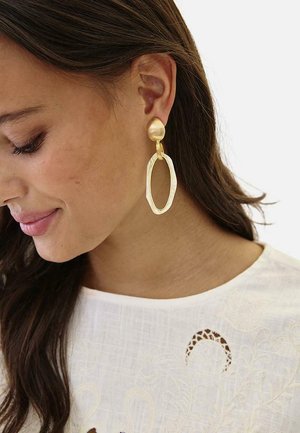 Mujer con cabello largo y castaño lleva pendientes grandes de aro dorados irregulares y una blusa blanca texturizada con detalles de encaje, mirando hacia abajo con suavidad.