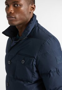 Giubbotto imbottito blu navy con colletto alto, chiusura a doppio petto con bottoni, tasca frontale e finitura testurizzata. Patch logo sulla manica.