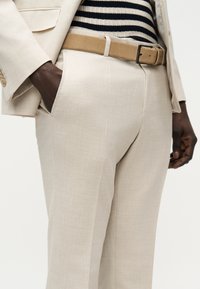 Personne portant un pantalon et une veste de costume beige avec une ceinture marron clair et un haut rayé, les mains près des poches sur un fond neutre.