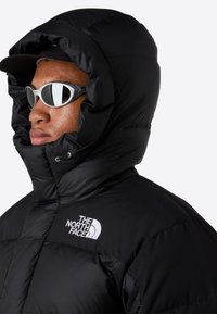 Puffjakke i sort med en høj, isoleret hætte og det hvide North Face-logo på brystet. Modellen bærer solbriller.