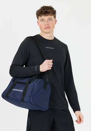 Nuori mies pukeutuneena mustiin urheiluvaatteisiin ja kantamassa tummansinistä duffel-laukkua, jossa on "ENDURANCE"-logo, olkapäällä.