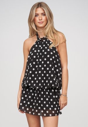 Donna con lunghi capelli biondi che indossa un vestito nero senza maniche con pois bianchi, in piedi davanti a uno sfondo semplice.