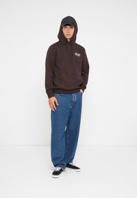 Bruine hoodie met een borstzak, gecombineerd met losse blauwe jeans en zwarte sneakers. De pet is zwart met een gebogen rand.