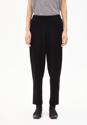 Personne portant un pantalon noir droit, une chemise à manches courtes rayée et des bottines noires à bout carré, se tenant devant un fond blanc.