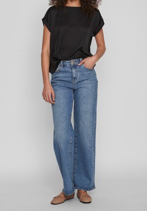 Femme portant un haut noir ample à manches courtes, un jean bleu taille haute à jambes larges, et des ballerines beige, debout devant un fond gris.