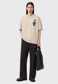 Camiseta de algodón beige con un gráfico de calavera negra, combinada con pantalones negros sueltos y un bolso negro texturizado con un logo.