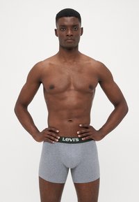 Moški brez srajce stoji s hands on hips, oblečen v sive Levi's bokserice z črnim pasom na enobarvni beli podlagi.