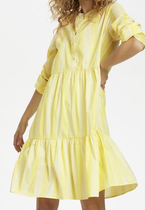 Femme portant une robe ample rayée verticalement jaune et blanche avec manches longues et un ourlet à volants, debout avec une main sur la hanche.