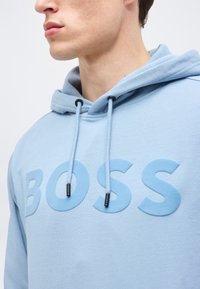 Jeune homme portant un sweat à capuche bleu clair avec l'inscription BOSS en relief sur la poitrine et des cordons pendants de la capuche.