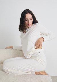Ensemble de lounge blanc comprenant un haut à manches longues avec un logo "BOSS" noir et un pantalon avec taille élastique. Tissu doux avec une texture lisse.