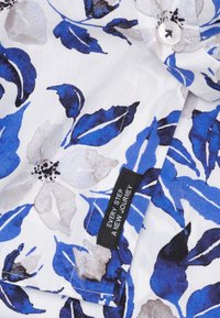 Bloemritshirt met een blauw-wit patroon met grote bloemen en bladeren. De stof is lichtgewicht en heeft een klein label met de tekst "IEDERE STAP EEN NIEUWE REIS."