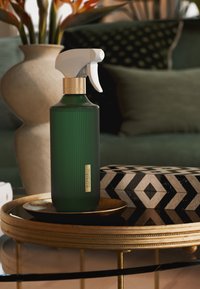 Bottiglia spray in vetro verde con texture rigata, ugello bianco e dettaglio dorato. Posizionata su un vassoio rotondo dorato con una scatola decorata.