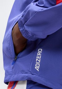 Χέρι στην τσέπη ενός μπλε μπουφάν με ελαστικό μανσέτα και φερμουάρ, με λευκό κείμενο "ADIZERO" στο κάτω μανίκι.