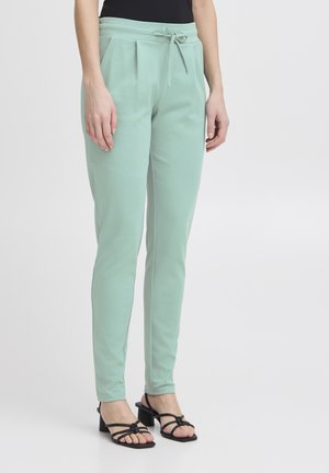 Mintgroene taps toegesneden broeken met een trekkoord in de taille, plooien aan de voorkant en een zachte textuur, gecombineerd met zwarte haksandalen.