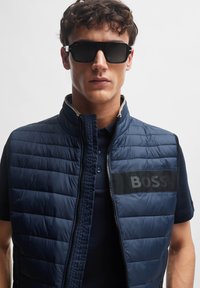 Námornícky puffový vest s prešívanou textúrou, s predným zipsom a čiernym logom "BOSS" na hrudi. Skombinovaný s čiernymi slnečnými okuliarmi, nosený cez námornícky polo.